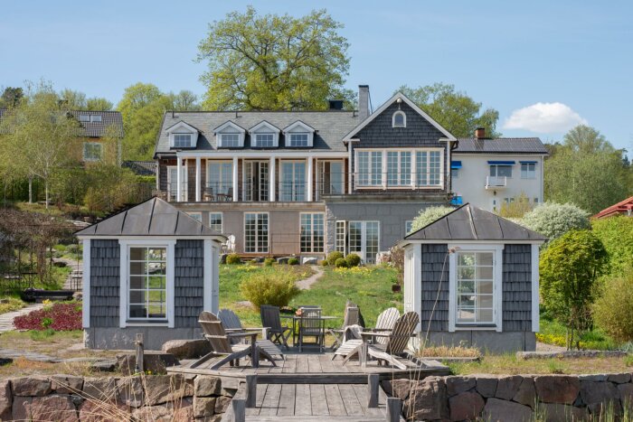 Stort hus med två små uthus och en träbrygga, omgiven av trädgård och grönska under en klarblå himmel.