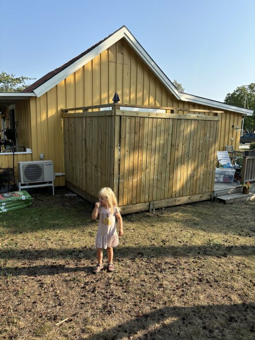 En blond flicka står framför en nybyggd altan med trästaket och ett gult hus i bakgrunden.