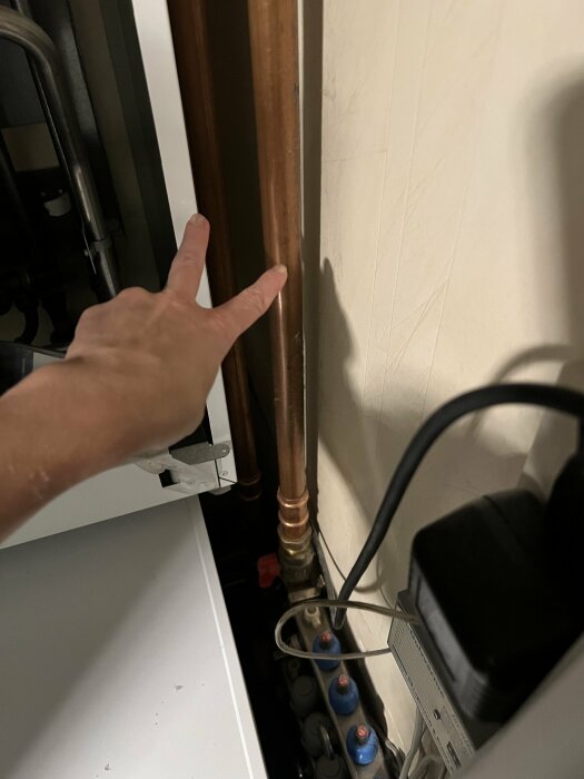 En hand pekar på två kopparrör med vattenanslutningar, omgiven av en elkabel och elektronisk apparatur, fotad i en teknisk installation.