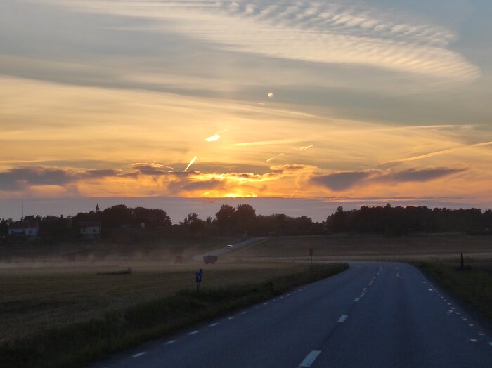 Solnedgång över en landsväg med fält på båda sidor; damm från en skördetröska syns nära marken och färgar himlen i gyllene nyanser.