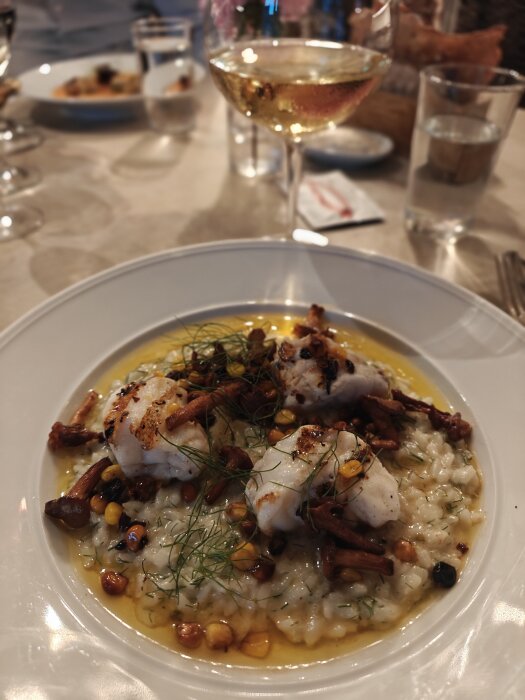 En tallrik med risotto, garnerad med kantareller och grillad marulk, serverad på ett dukat bord med ett glas vin och vatten i bakgrunden.