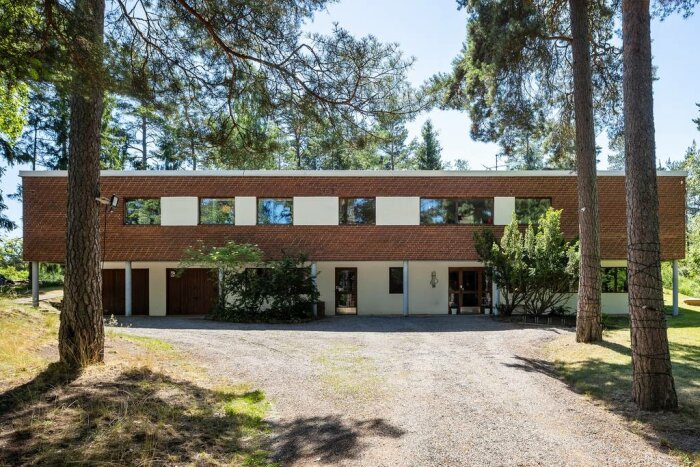 Modern villa med platt tak och tegelväggar omgiven av träd. Huset har stora fönster och en grusad infart. Skogvaktarvägen 22, som visas från framsidan.