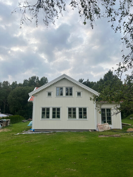 Vit byggnad med snickarglädje på nock, omgiven av träd och gräsmatta under molnig himmel. En lastpall och byggmaterial ligger vid sidan av huset.