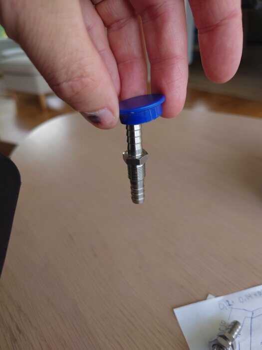 En genomföring med blå plastkåpa och metallskruv hålls upp med fingrar ovanför ett bord.