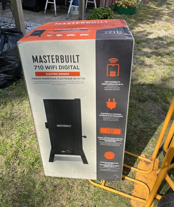 Kartong med en Masterbuilt 710 WiFi Digital Electric Smoker står på gräsmattan, redo att packas upp.