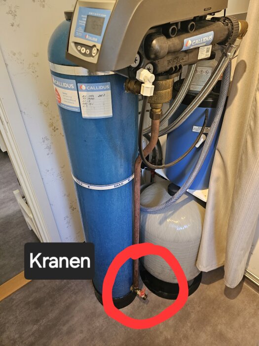 Bild av ett Callidus vattenfilter med fokus på en avrinningskran i botten, som är markerad med en röd cirkel och texten "Kranen".