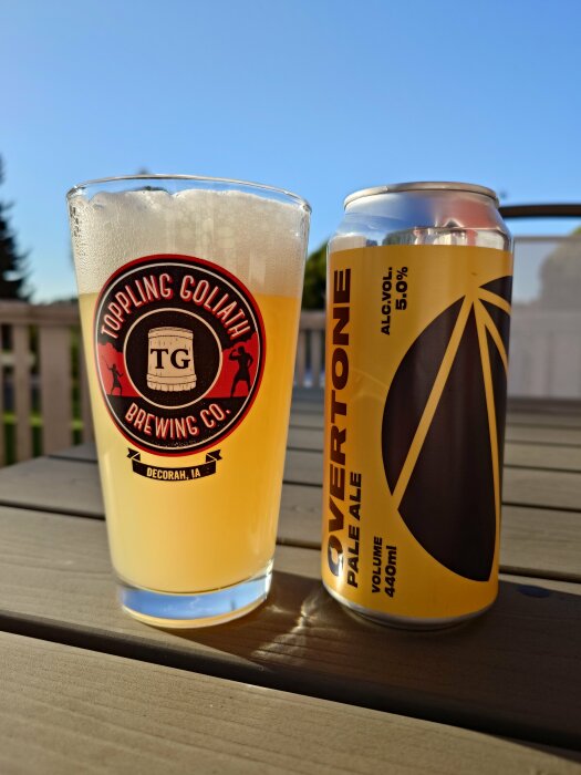 Ett glas med ljus öl från Toppling Goliath Brewing Co. och en burk Overtone Pale Ale på ett trädäck i solsken.