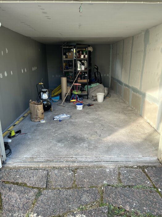 Garage interiör med grå väggar under renovering; hyllor med verktyg och material, oavslutat golv med verktyg och material spridda på ytan.