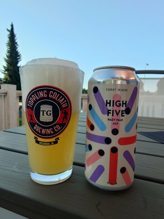 En burk Fuerst Wiacek High Five Hazy Pale Ale och ett ölglas med Toppling Goliath Brewing Co-logga, fyllt med pale ale, placerade på ett bord utomhus.