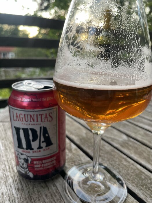 Lagunitas IPA ölburk på ett träbord med ett halvfullt glas öl bredvid, mot en suddig bakgrund av träd och staket.