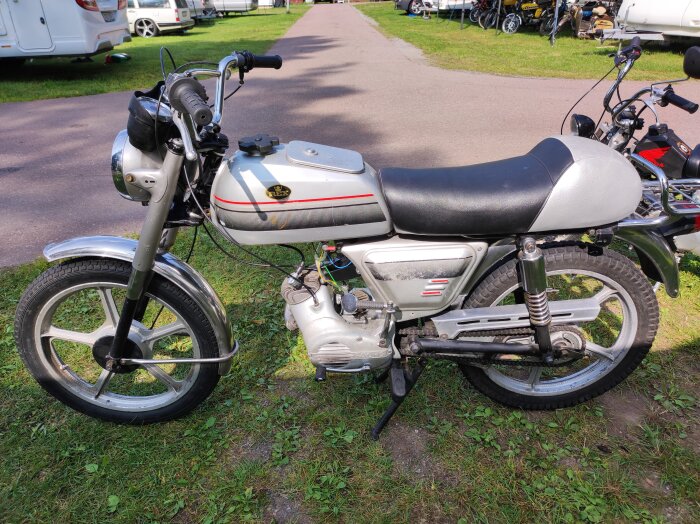 Silverfärgad REX Sprint-moped parkerad på gräs, med asfaltväg och andra fordon i bakgrunden.