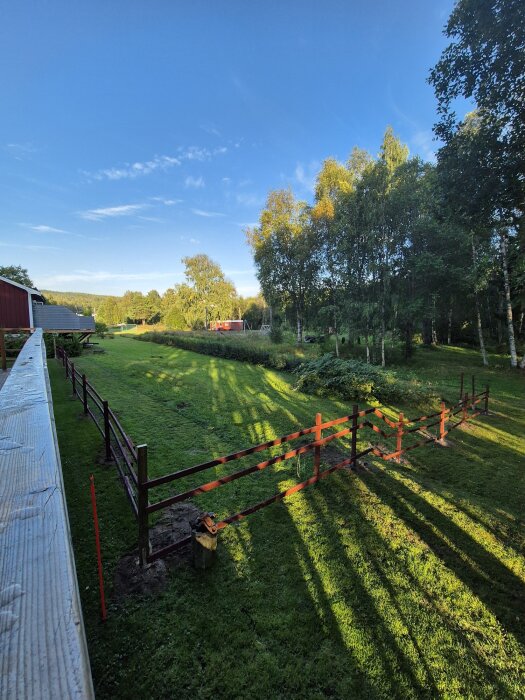 Staket med två träbalkar över en grön gräsmatta, omgiven av träd, som hindrar tillgång till ett garage i ett bostadsområde.