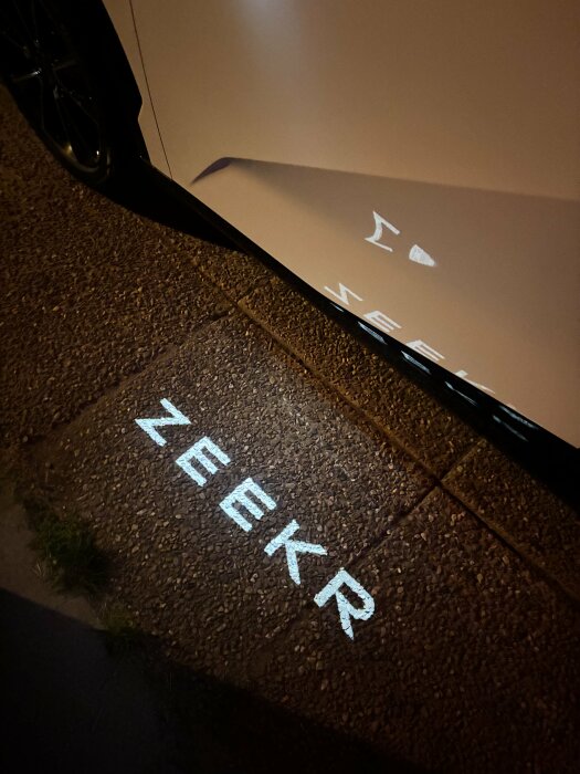 Bilens dörrprojiceringsljus visar texten "ZEEKR" på trottoaren bredvid bilen under kvällen.