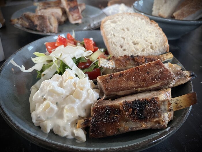 Grillad fårbringa serverad på en tallrik med potatissallad, skivad baguette och sallad med tomater. Totalt två tallrikar syns, en i bakgrunden och en i förgrunden.