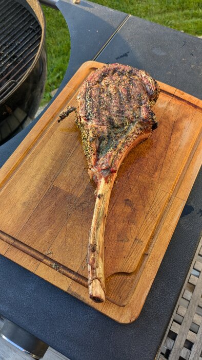 En grillad tomahawkstek ligger på en skärbräda utomhus bredvid en kolgrill.