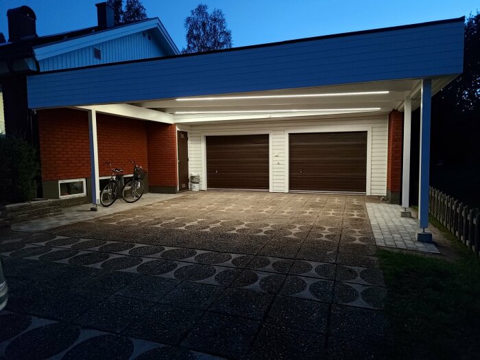 Utsikt över uppfart och carport med två cyklar, belysning från ledstrips på taket och två garageportar i mörkt trä, mot en husfasad av röd tegel och vit panel.