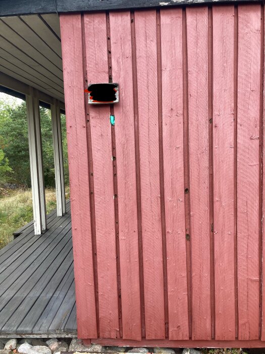 Röd träfasad med hål av olika storlekar samt en öppen lucka ovanför, som visar isolering bakom ytterpanelen på ett sommarhus från 70-talet.