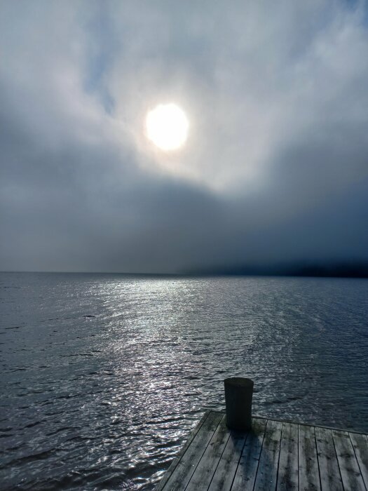 Solig morgon vid en brygga med utsikt över en lugn sjö. Solen går upp över horisonten och sprider sitt ljus genom disiga moln, som reflekteras i vattnet.