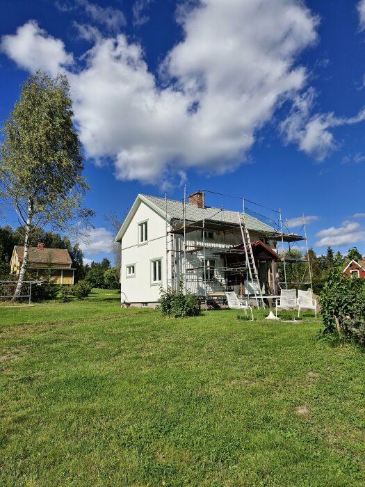 Hus med byggnadsställningar på gräsmatta under en blå himmel med några vita moln, förberedelser för renovering pågår.