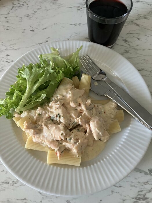 En tallrik med rigatoni-pasta och laxsås, toppad med sallad bredvid bestick, samt ett glas röd dryck i bakgrunden.