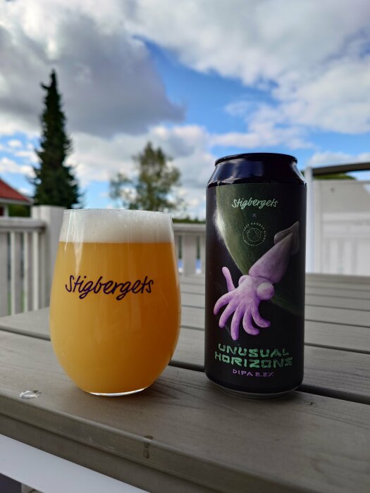 En burk och ett glas av Stigbergets "Unusual Horizons" DIPA, med juicig färg, på ett bord utomhus med en molnig himmel i bakgrunden.