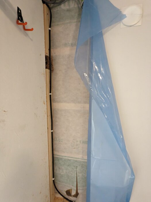 Fastighetsvägg med exponerad installationskabel som saknar spikskydd, fixerat med vita kabelband. Blå plastfolie delvis avlägsnad, visar isolering bakom.