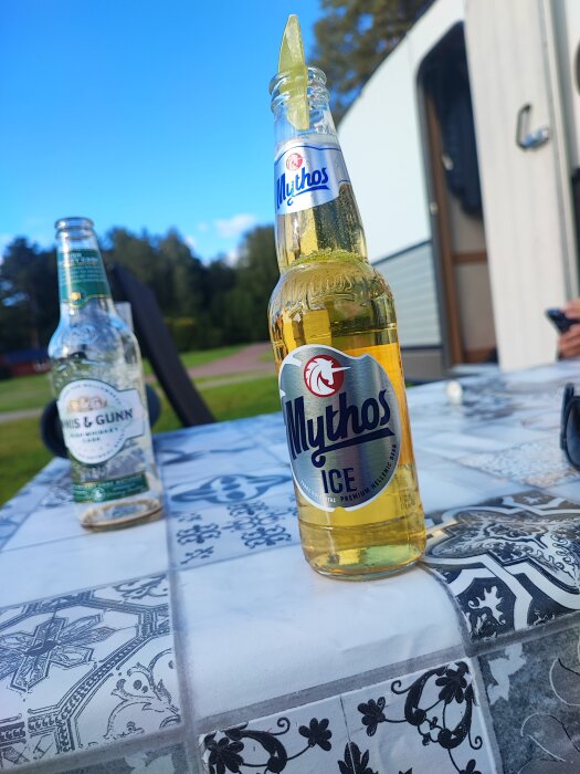 En Mythos Ice flaska med lime i öppningen står på ett kaklat bord utomhus, med en tom Innis & Gunn flaska i bakgrunden.