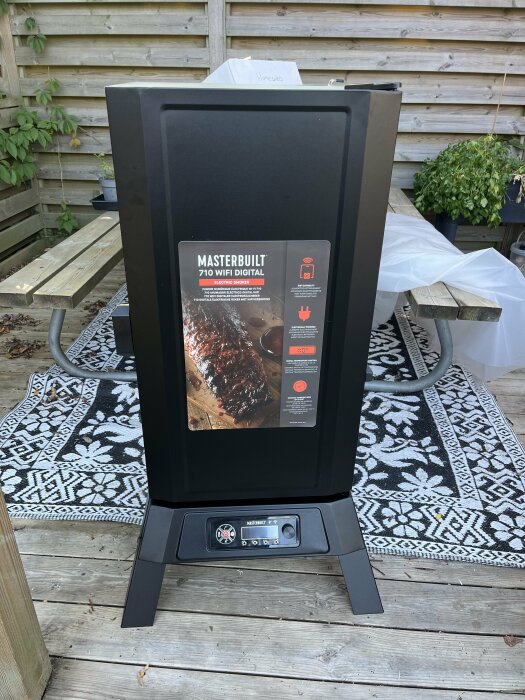 Masterbuilt 710 WiFi Digital Electric smoker monterad och redo att användas, placerad på en altan framför ett picknickbord med växter i bakgrunden.