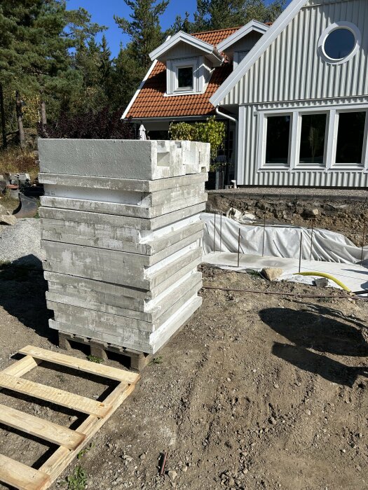 Staplade betongblock på en byggarbetsplats framför ett hus med rött tegeltak och vita träpaneler.