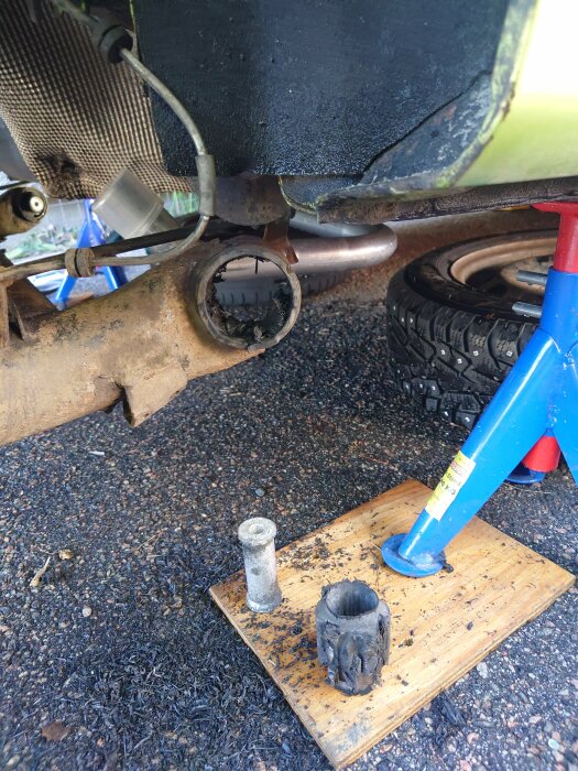 Bil på pallbock med rostig kopplingsdel borttagen, smutsiga bildelar på trädplatta under, förberedelser för reparation.