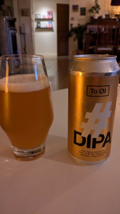 En ölburk märkt "#DIPA" från To Øl och ett halvtom ölglas fyllt med gyllene öl står på ett bord i ett belyst vardagsrum.