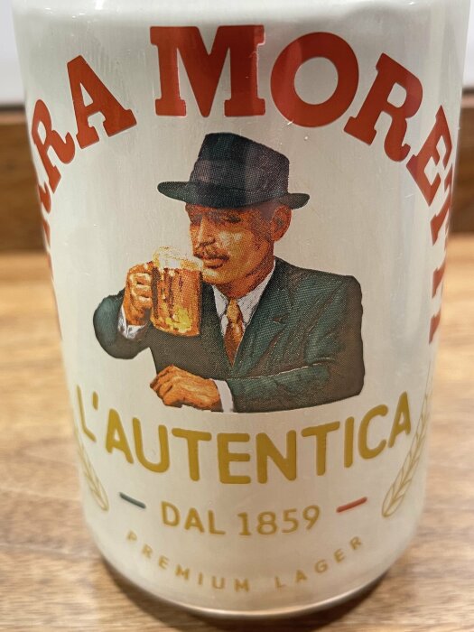 En närbild av en Birra Moretti beer can, med en illustration av en man i kostym som håller ett ölglas. Texten på burken lyder "L'Autentica", "Dal 1859".