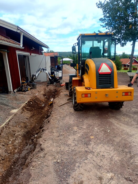 Gul fordon och grävmaskin vid en utgrävd diken framför en röd byggnad, för markisolering och byte av vägtrumma.