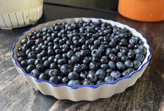 En pajform fylld med blåbär står på en mörk arbetsyta, redo för att bakas till blåbärspaj enligt recept.