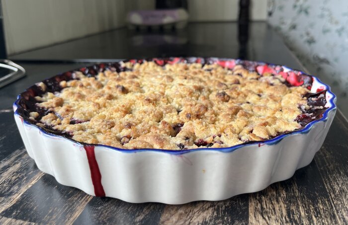 Blåbärssmulpaj i en vit pajform med en gyllene, smulig yta och blåbärssaft som rinner över kanten, placerad på en mörk träyta.