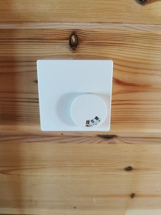 Vit termostat monterad på en vägg i träpanel, med en ratt som visar temperaturinställningarna 10, 15 och 20 grader Celsius i svart text.