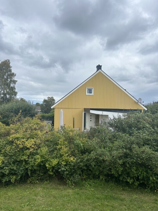 Bild på en gulmålad enplansvilla med stor taklutning, omgiven av lummiga buskar och träd, under en mulen himmel.