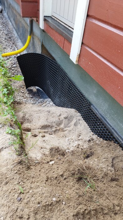 Bild av husgrund med delvis avlägsnad jord, en fastmonterad Platonmatta, exponerade dräneringsrör och förberedd yta för återfyllning av grus och sand.