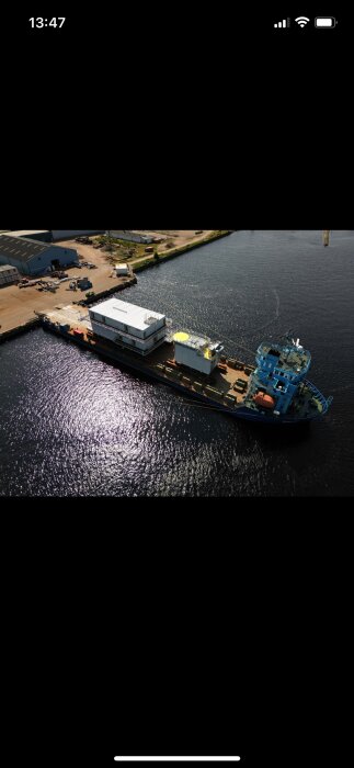 Vindkraftsplattform med två gråa moduler placerade på ett fartyg i en hamn, omgivna av vatten och byggnader.