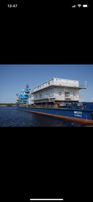 Vit modul till vindkraftsplattform på ett blått fartyg med texten "MERI TURKU" på sidan, förankrad i en hamn under klar himmel.