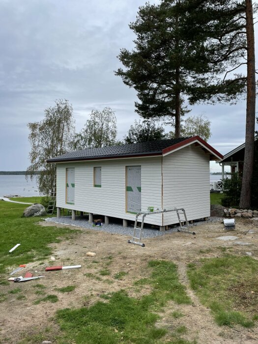 Småstuga under byggnation nära en sjö. Plåttak och träpanel är på plats, men nockplåt och gavelbeslag saknas. Dörrar och fönster ska installeras och målas rött.