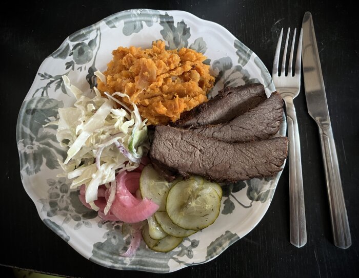 En tallrik med rökt brisket, potatismos av sötpotatis, coleslaw med rödlök, picklad rödlök och inlagd gurka. Bestick ligger bredvid tallriken.