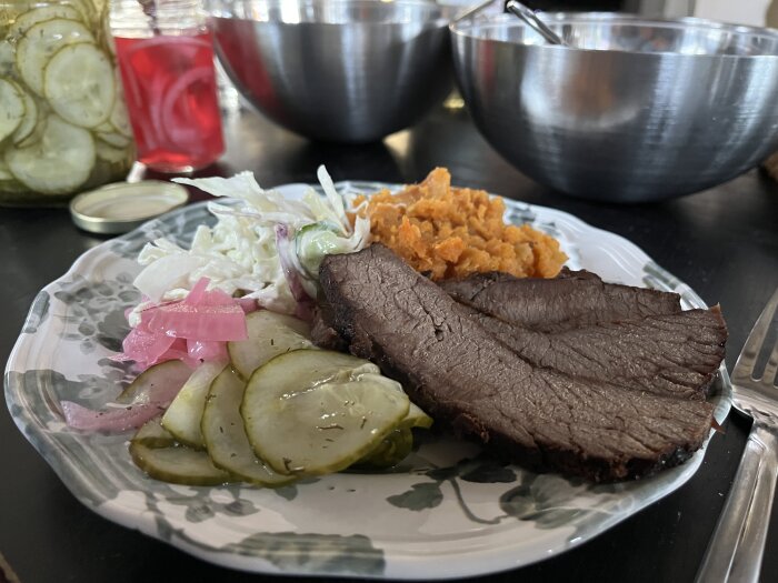 En tallrik med rökt brisket, potatismos, coleslaw, inlagd gurka och picklad rödlök på ett bord. I bakgrunden syns kärl och burkar med inläggningar.