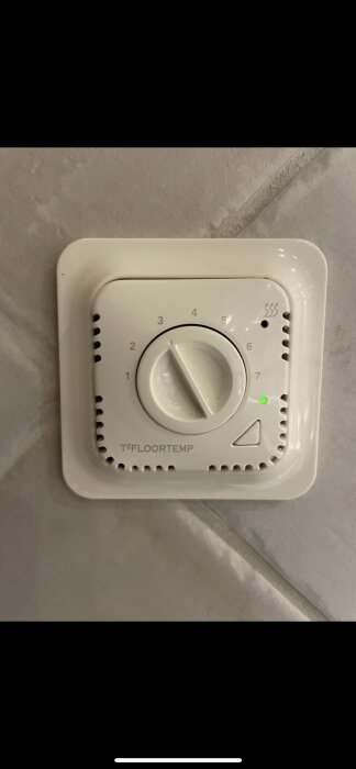 Vit T2Floortemp termostat installerad på en vägg, med kontroll för att justera temperaturen mellan nivå 1 till 7 och en tänd grön indikatorlampa.