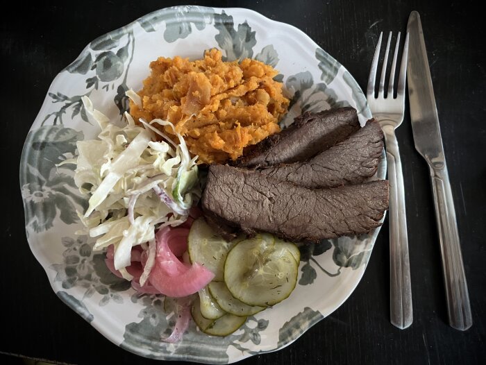 En tallrik med skivad brisket, picklad rödlök, inlagd gurka, coleslaw och sötpotatismos, med kniv och gaffel bredvid.