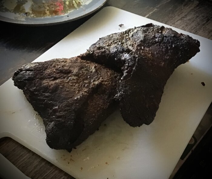 Två halvor av en rökt brisket ligger på en vit skärbräda, med en tallrik mat suddigt i bakgrunden.