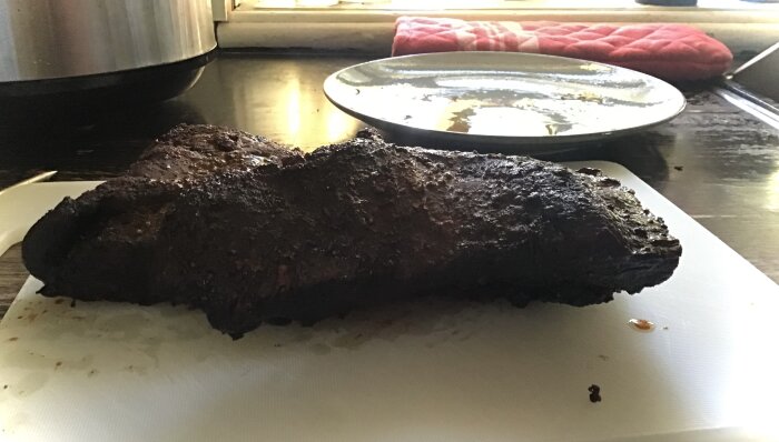 Nyutplockad brisket från röken ligger på en skärbräda och är redo att skäras upp för servering. En tallrik och en kökshandduk syns i bakgrunden.