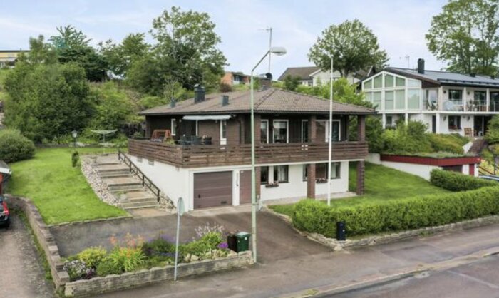 En sutterängvilla med trädgård, stenbelagda trappsteg och garage. Huset ligger nära en trafikerad väg och omges av grönska och andra bostäder.