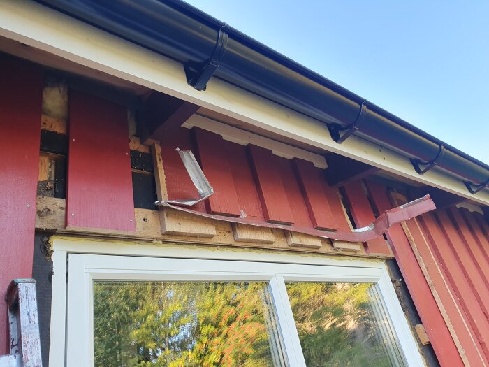 Byggprojekt: byte av fasadpanel där nya röda paneler monteras ovanför ett vitt fönster med reflekterande glas, takfot och överbleck synliga.