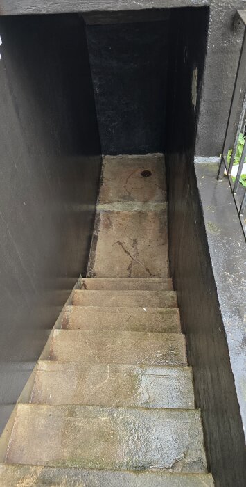 Betongtrappa ned till källare med igensatt dörr och fuktfläckar på väggarna; avlopp synligt på källargolv.